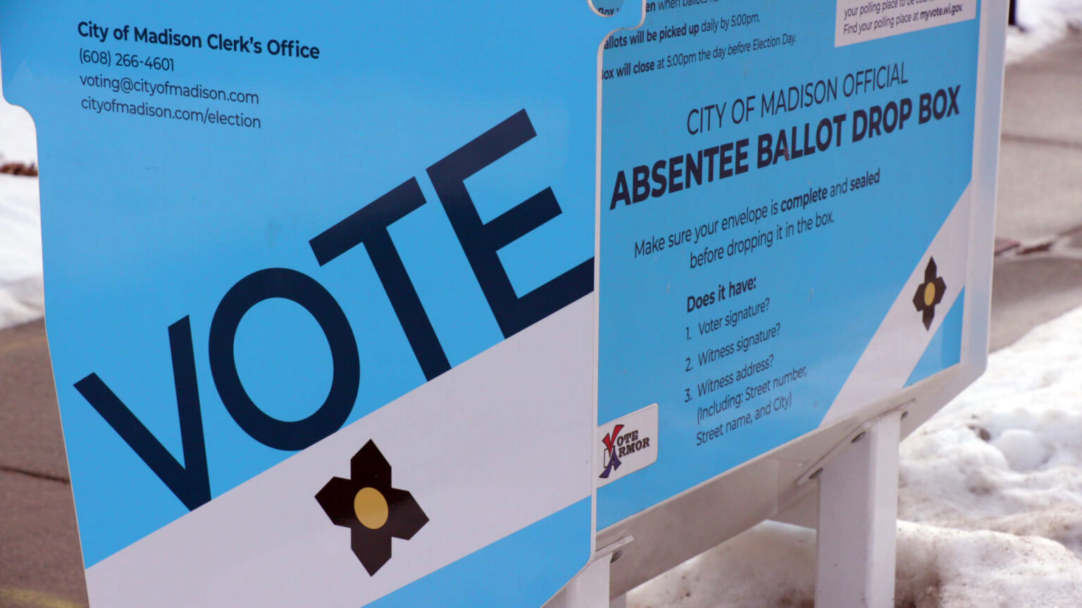 A metal box absentee ballot drop box that includes the words City of Madison Clerk's Office with a phone number, email address and URL on one side and additional words on another side stands next to a sidewalk amid snowdrifts.