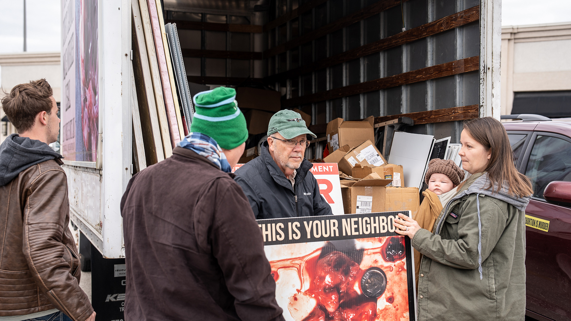 Matthew Trewhella holds a printed sign with the words "This Is Your Neighbor" and a photo of bloody tissue and a U.S. dime while standing among several other people, one of whom is holding an infant, in front of the back entrance to the cargo area of a moving truck containing other signs and cardboard boxes.