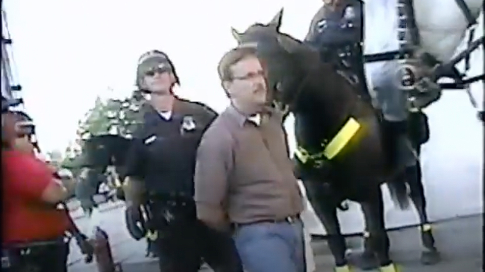 A low resolution screenshot from a video showing Matthew Trewhella standing with his arms behind his back and being led by a police officer, with the horses of two mounted police to the side.