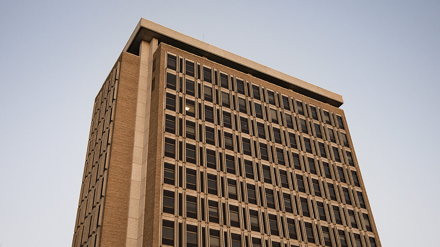 A tower with a brick-and-concrete surface framing multiple stories with four windows on one face and 12 on another face stands under a cloudless sky, with sunshine illuminating one side of the building.