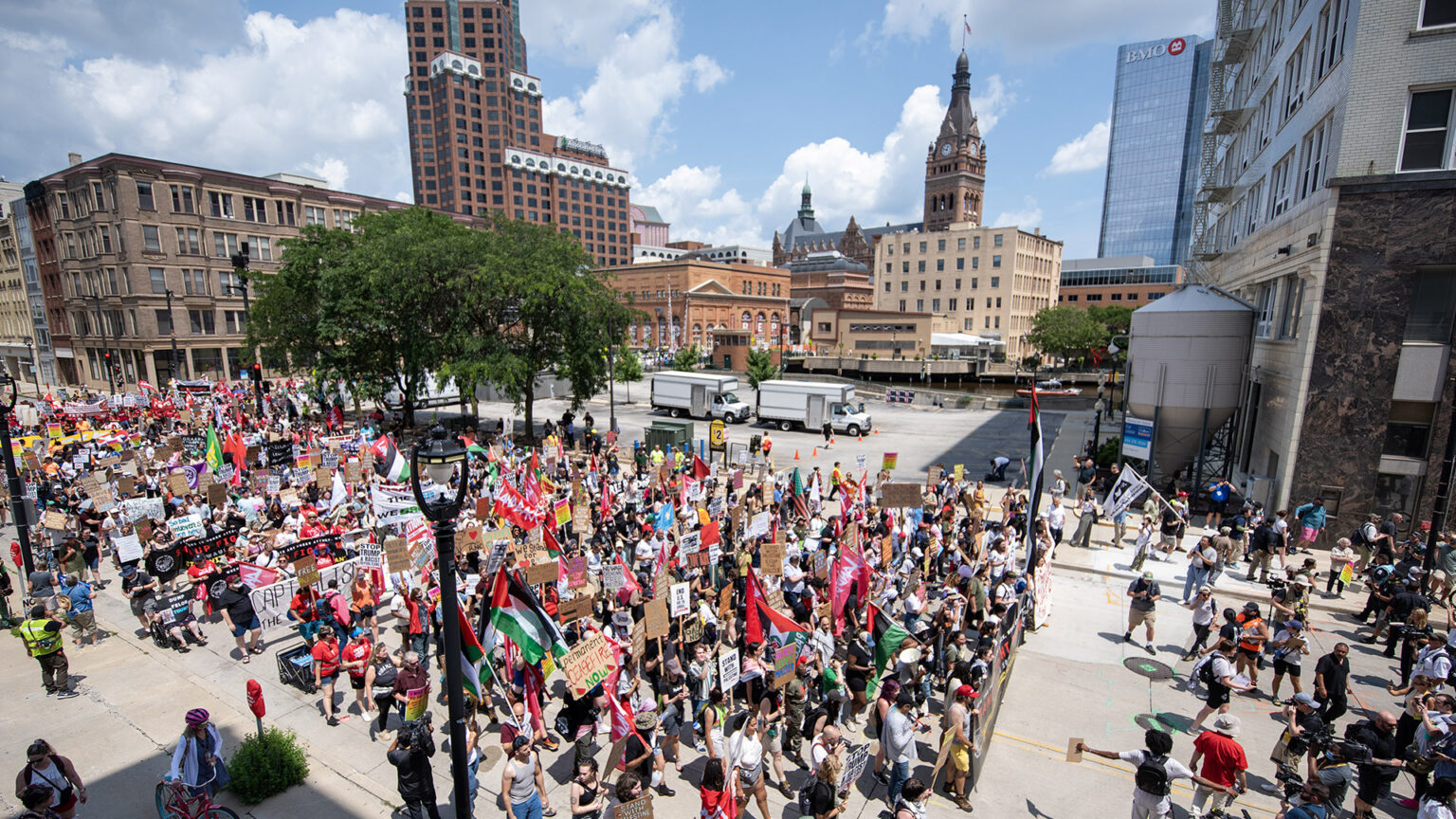 Protesters carrying preprinted signs, banners on poles and flags march along a city street, with trees, parking lots and multiple buildings of different architectural styles and heights in the background.