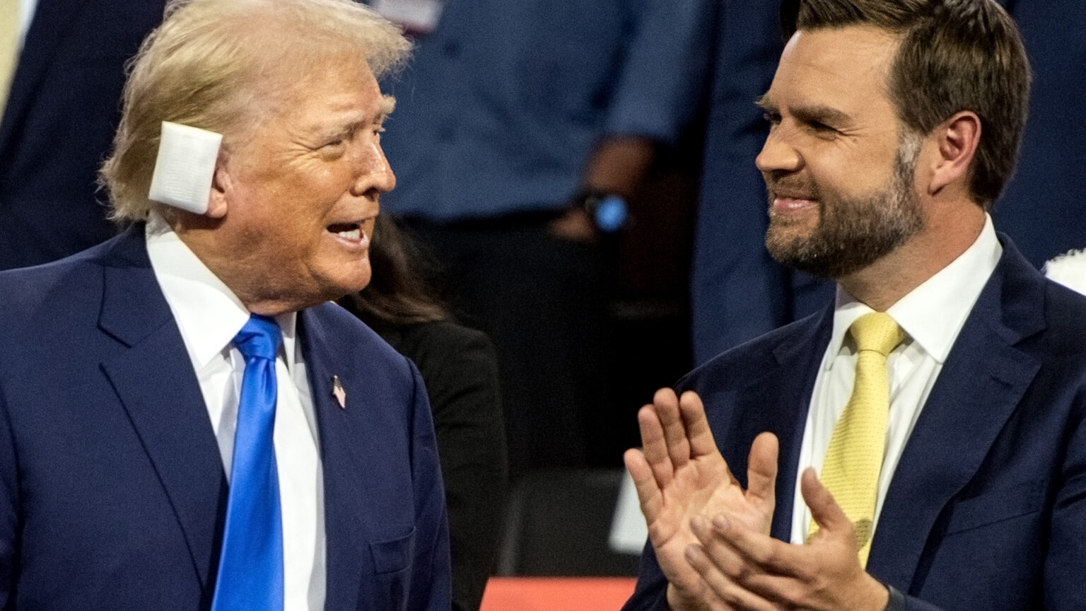 Donald Trump, who has a bandage over his right ear, speaks while facing JD Vance, who is clapping his hands.