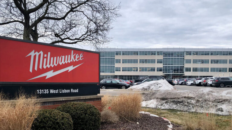 A sign with the Milwaukee Tool wordmark and lightning bolt logo is surrounded by bushes near the entrance to a parking lot, with a tree, parked cars and a multi-story office building in the background.