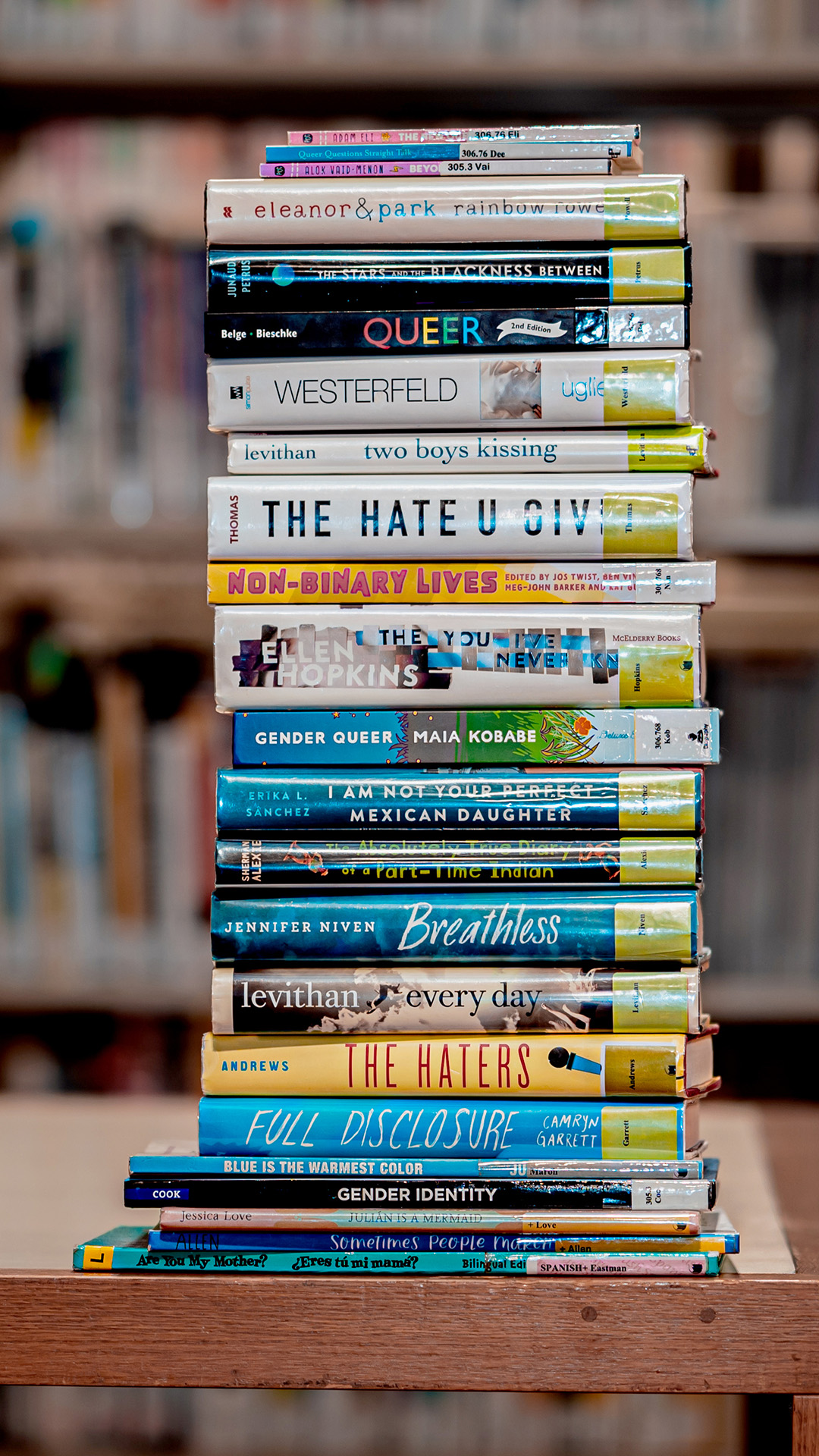 A stack of about two dozen books with transparent protective dust jacket covers and library labels stands on a wood table, with out-of-focus bookshelves in the background.