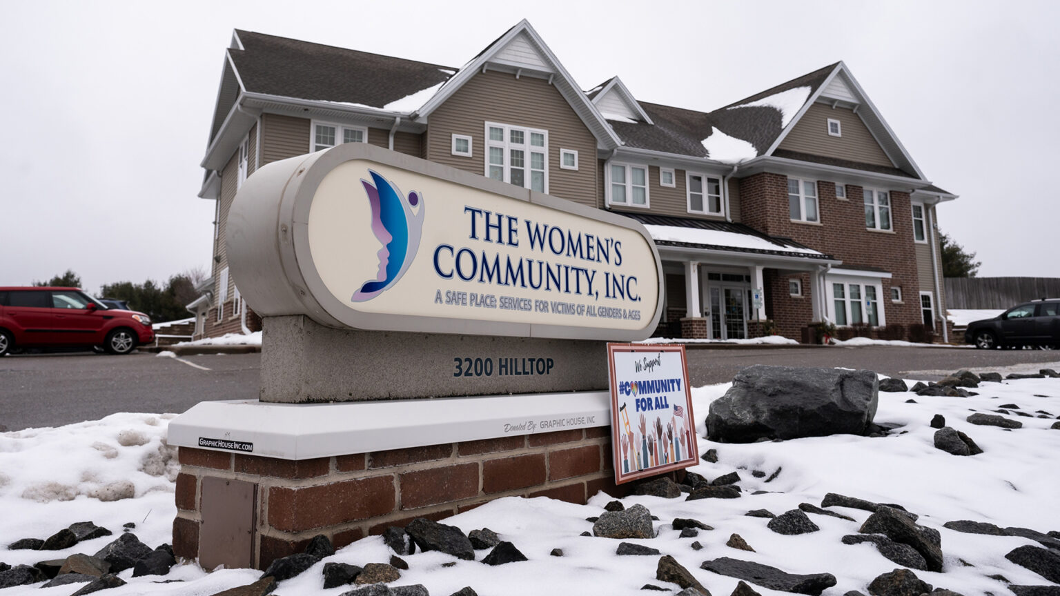 An unlit electrified oval sign with a logo of multiple faces and the words The Women's Community, Inc. and A Safe Place: Services for Victives of All Genders & Ages stands on a low brick platform in a snow-covered area of landscaping rocks in front of a driveway to a multi-story building with multiple gables and vehicles parked on multiple sides.