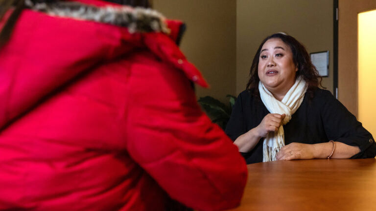 Monica Lo sits at a wood table and speaks while facing another person in the foreground, in a room with a glass-windowed door in the background.