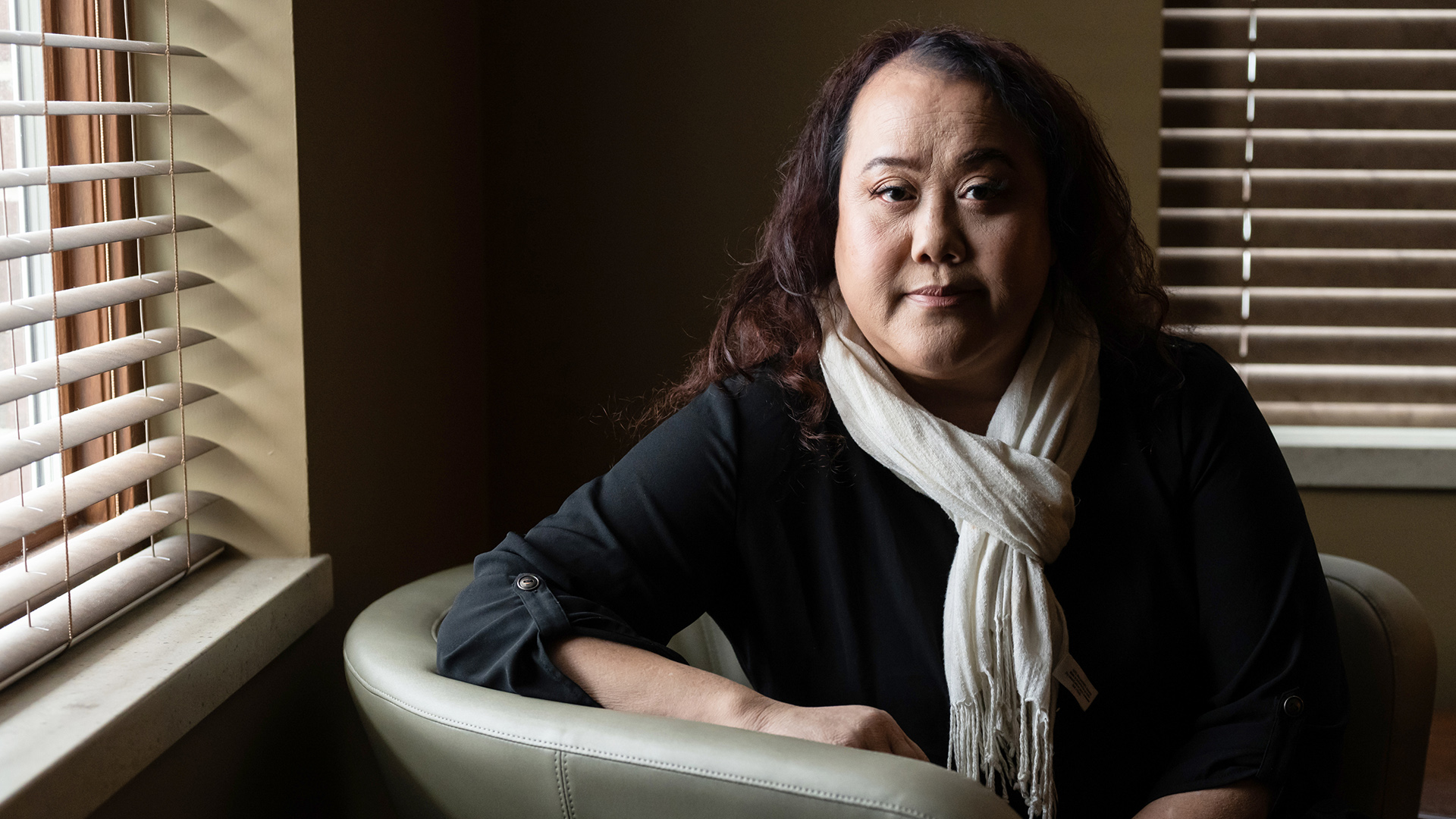 Monica Lo poses for a portrait while seated in a leather chair in the corner of a room with open and closed blinds covering windows.