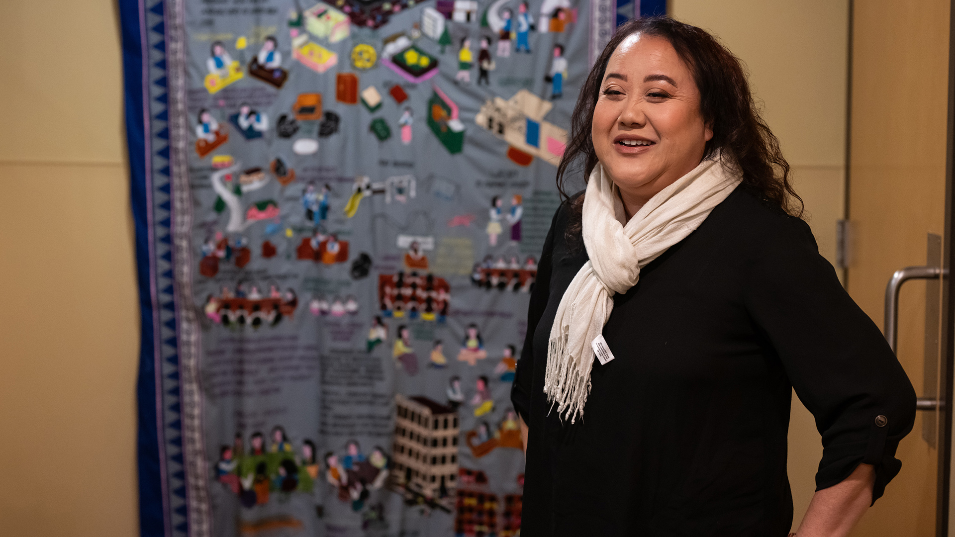 Monica Lo stands and speaks in a room with a Hmong story cloth displayed on a wall in the background.