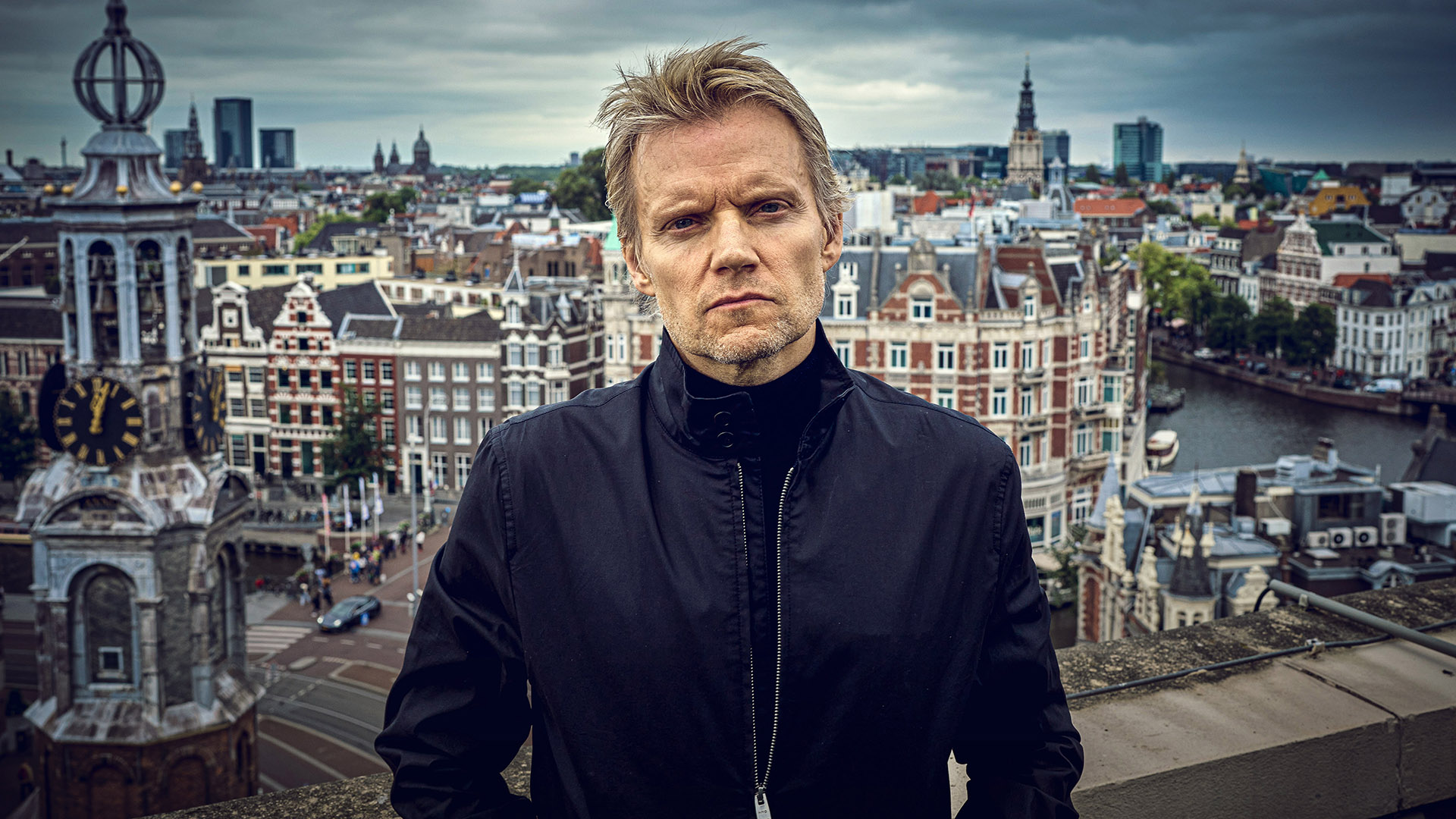 A man in a black jacket and serious expression stares at the camera with the city of Amsterdam in the background.