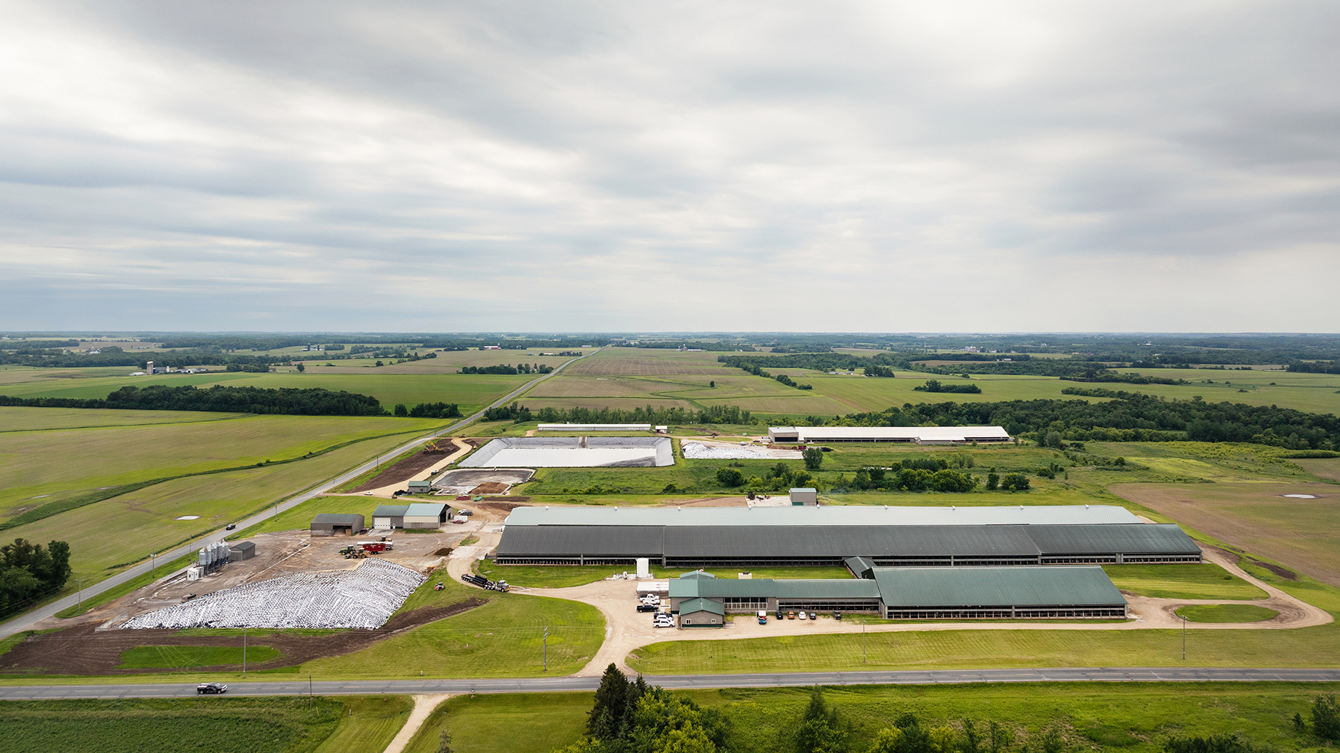 An aerial photo shows multiple large barns and other buildings along with a holding pond and gravel driveways at the intersection of two paved roads, with farm fields and wooded areas stretching to the horizon under a cloudy sky.