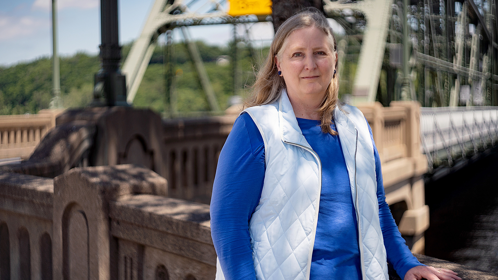 Kim Dupre poses for a portrait while standing next to a stone balustrade at the entrance to a metal truss bridge above a river, with out-of-focus trees on the opposite shore in the background.