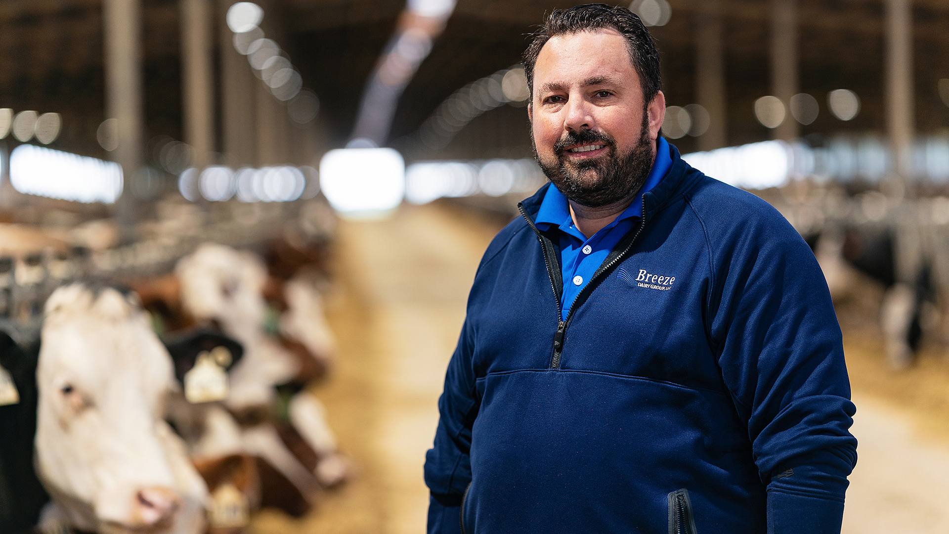 Gregg Wolf poses for a portrait while standing in a large barn, with out-of-focus cows standing in feeder panels in the background.