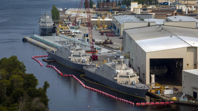 Two naval vessels surrounded by a row of floating buoys sit alongside a wharf with multiple metal-walled buildings, cranes and other construction equipment on shore, with another docked ship and buildings in the background, as well as trees on the opposite shore in the foreground.