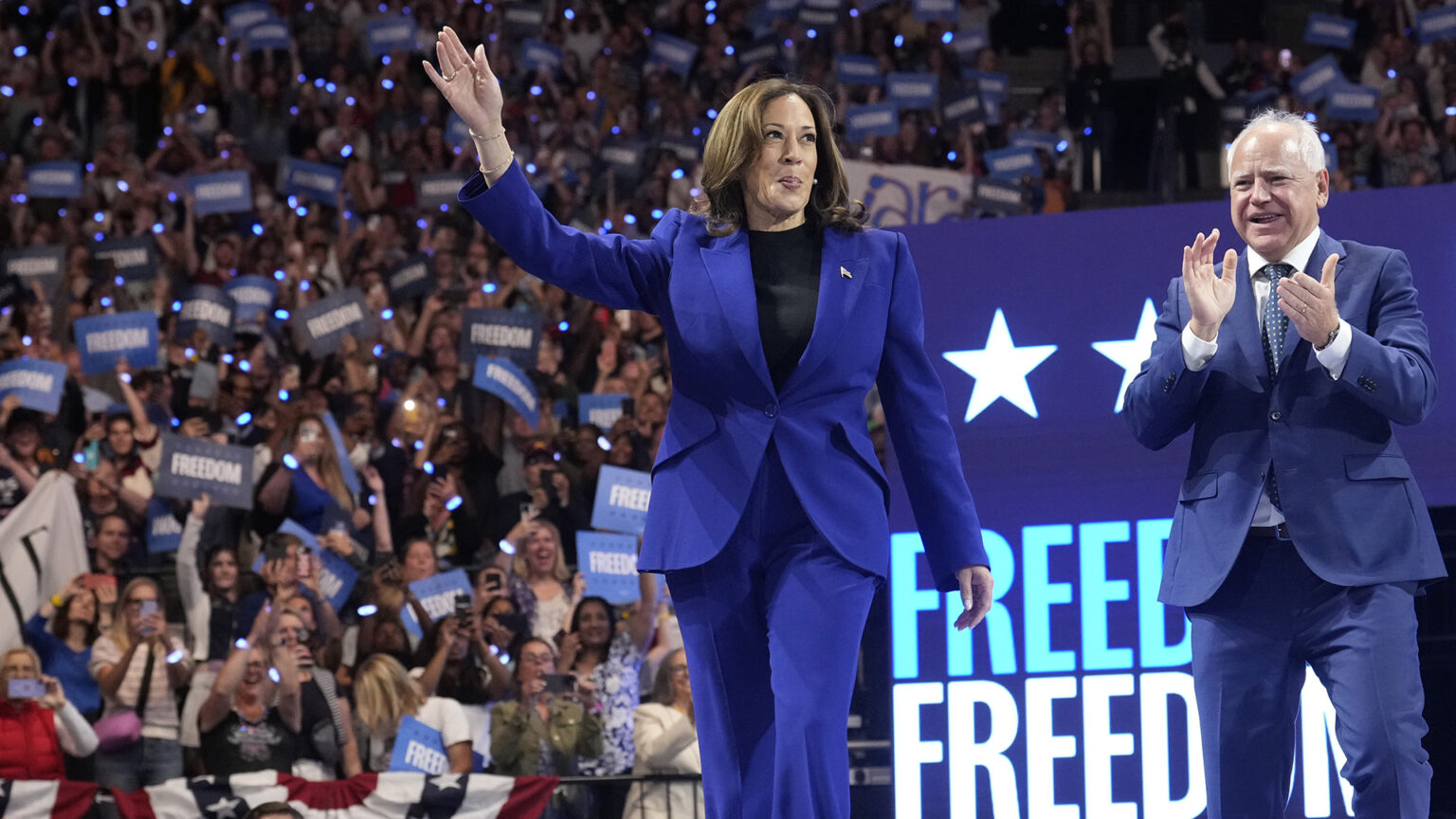 Kamala Harris stands on a stage and waves her right hand while Tim Walz stands to the side and claps, with an illuminated sign with the word Freedom and an audience of people holding up cell phone flashlights and political signs reading Freedom on arena bleachers in the background.