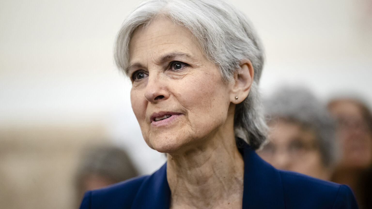 Jill Stein speaks while seated in a room with out-of-focus people seated behind her.