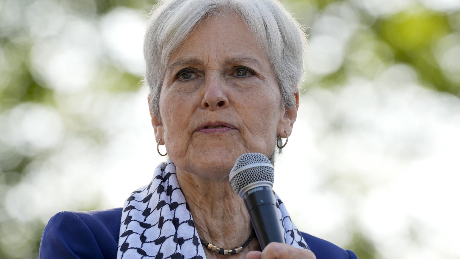 Jill Stein stands and speaks into a microphone, with out of focus tree foliage in the background.