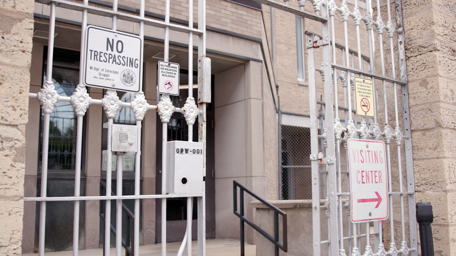 Multiple metal signs, including ones that state No Trespassing and Visiting Center with an arrow pointing right are affixed to painted wrought-iron gates attached to masonry wall that stands in front of a brick-and-masonry building with a short staircase leading to two doors with glass windows.