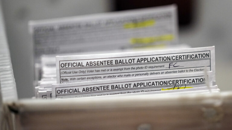 A stack of paper envelopes a label on their top edge that reads Official Absentee Ballot Application/Certification with a field filled by handwritten initials stand in the corner of a U.S Postal Service mail tub.