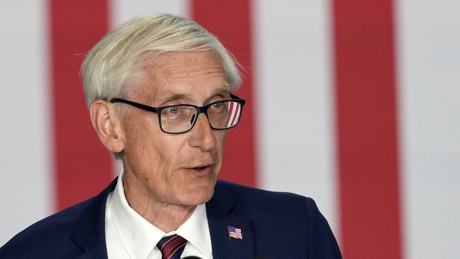 Tony Evers speaks while standing in front of the stripes of the U.S. flag in a vertical position.