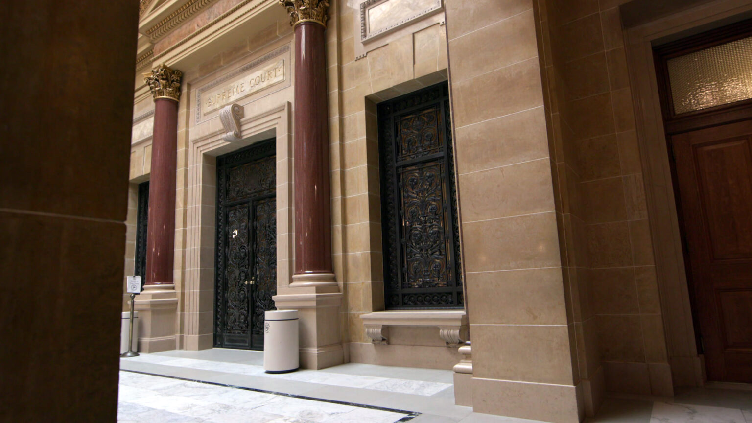 Double doors and windows to either side with wrought iron filigree are closed in a sunlit interior hallway of a building with marble masonry and two columns with Composite order capitals flanking the doorway.