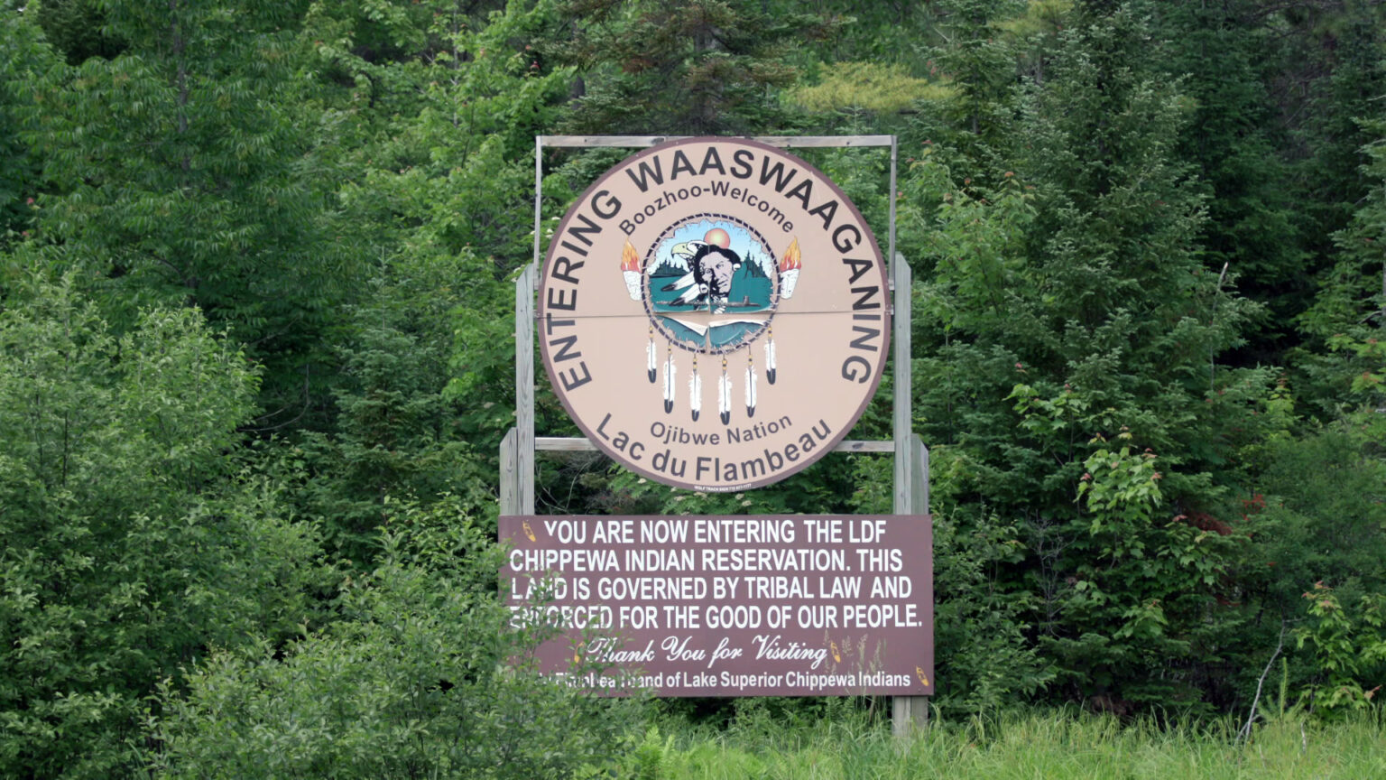 Two wood signs —  one reading Entering Waaswaaganing, Boozhoo-Welcome, Ojibwe Nation and Lac du Flambeau, surrounding the seal of the Lac du Flambeau Band of Lake Superior Chippewa, and another reading You are now entering the LDF Chippewa Indian Reservation. This land is governed by tribal law and enforced for the good of our people. Thank you for visiting. Fond du Lac Band of Lake Superior — are affixed to wood scaffolding in front of a wooded area.