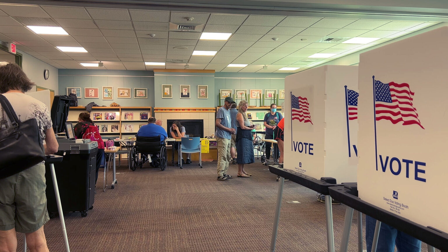 A person stands and faces a portable voting booth, with other booths comprised of a plastic table with metal legs and a privacy screen with U.S. flag graphics and the word VOTE on its sides, in a room with multiple people standing and seated at folding tables in the background, in a room with a carpeted floor and a fireplace on the back wall.