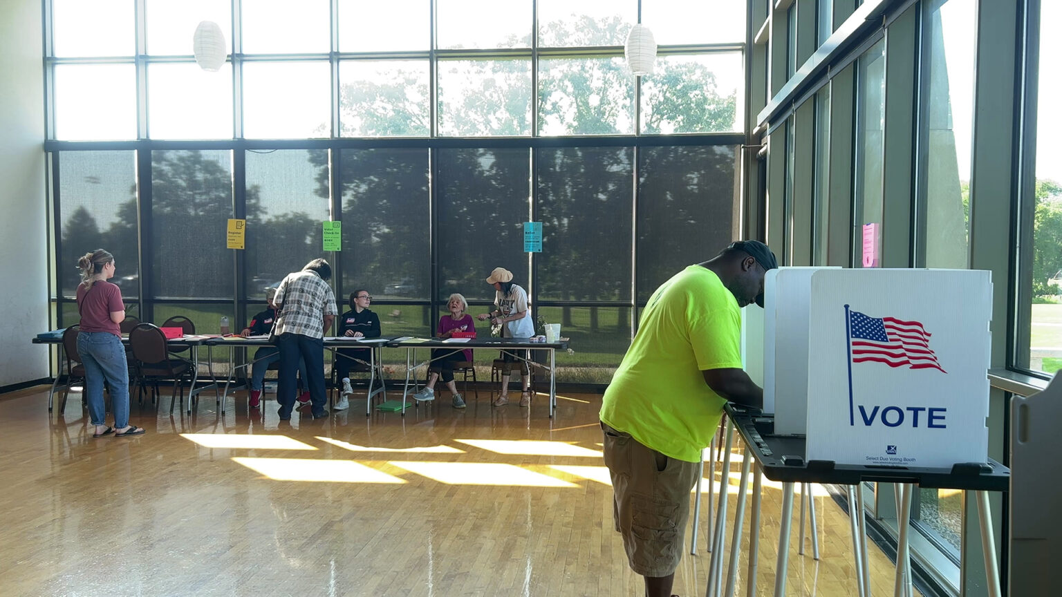 A voter stands and faces a portable voting booth comprised of a plastic table with metal legs and a privacy screen with a U.S. flag graphic and the word VOTE on its side, in a room with multiple people standing and seated at folding tables in the background, in a room with a sunlit polished wood floor and glass window walls, with trees and a lawn visible outside.