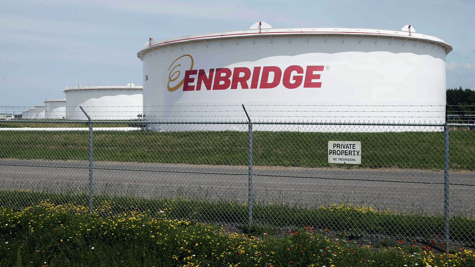 A chain link fence topped by barbed wire and a sign reading Private Property and No Trespassing stands in front of a gravel road and a row of industrial oil storage tanks with the Enbridge wordmark painted on the side of the one in front.