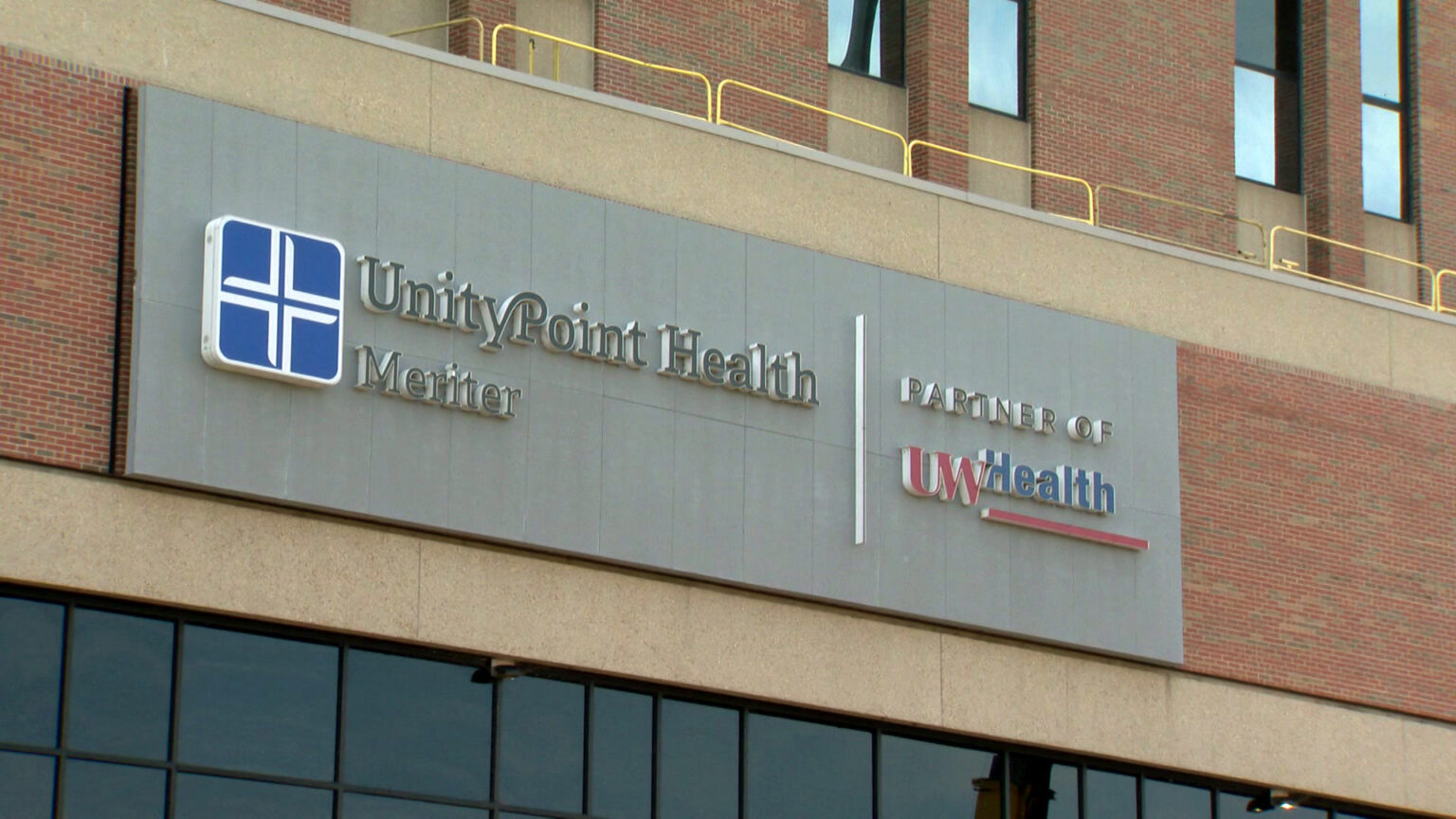 A metal building sign with the words Unity Point Health, Meriter and Partner of UWHealth is affixed to the extrior wall of a brick and concrete building with multiple tiers and window placements.