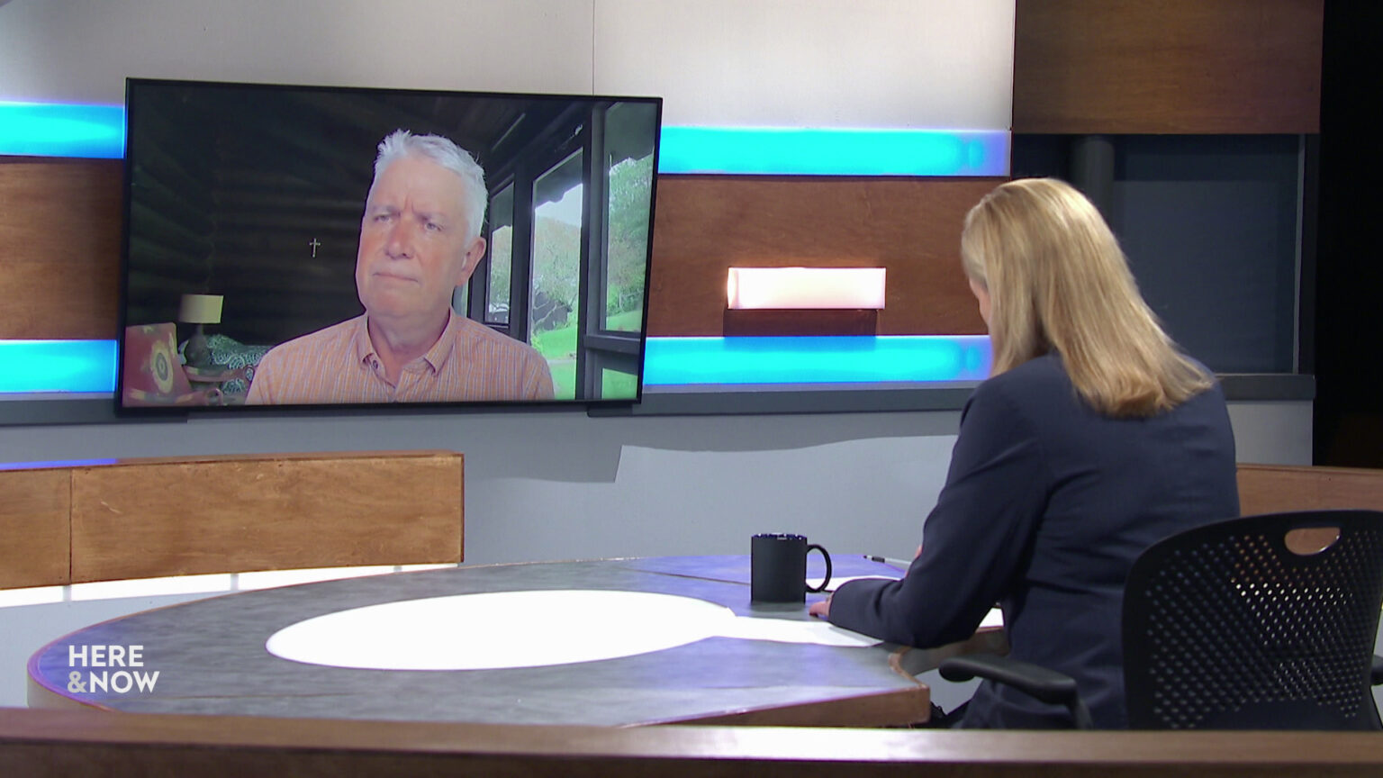 Frederica Freyberg sits at a desk on the Here & Now set and faces a video monitor showing an image of Bob Atwell.