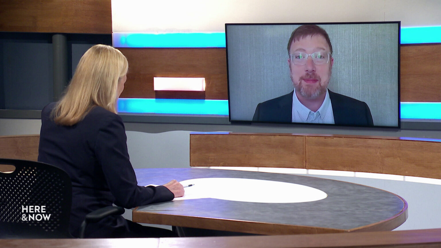 Frederica Freyberg sits at a desk on the Here & Now set and faces a video monitor showing an image of Ben Wikler.