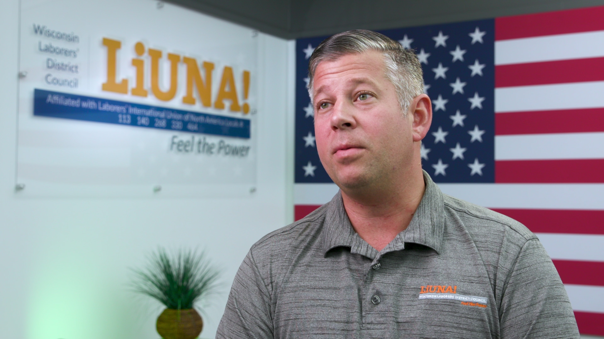 Kent Miller speaks with a U.S. flag, potted plant and sign reading 'Wisconsin Laborers District Council LiUNA!' in the background.