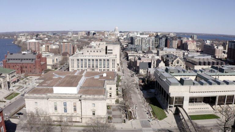 An aerial photo shows a cityscape of various buildings, the dome of the Wisconsin State Capitol and portions of two lakes visible in the background.