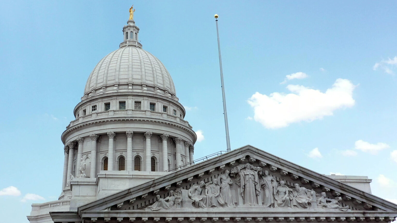 A flagpole is mounted to the top of one wing of a marble masonry building with Composite order columns supporting a pediment with relief statuary in front of a dome topped by a statue, under a partly cloudy sky.