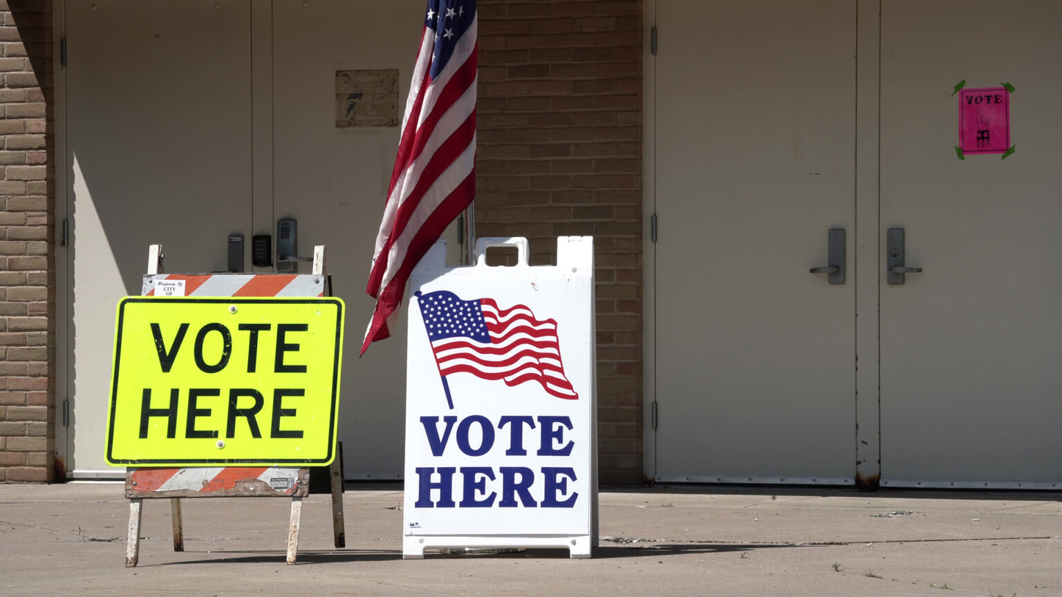 Two folding highway and sandwich board signs that read Vote Here stand on a concrete patio outside multiple double doors for a brick building.