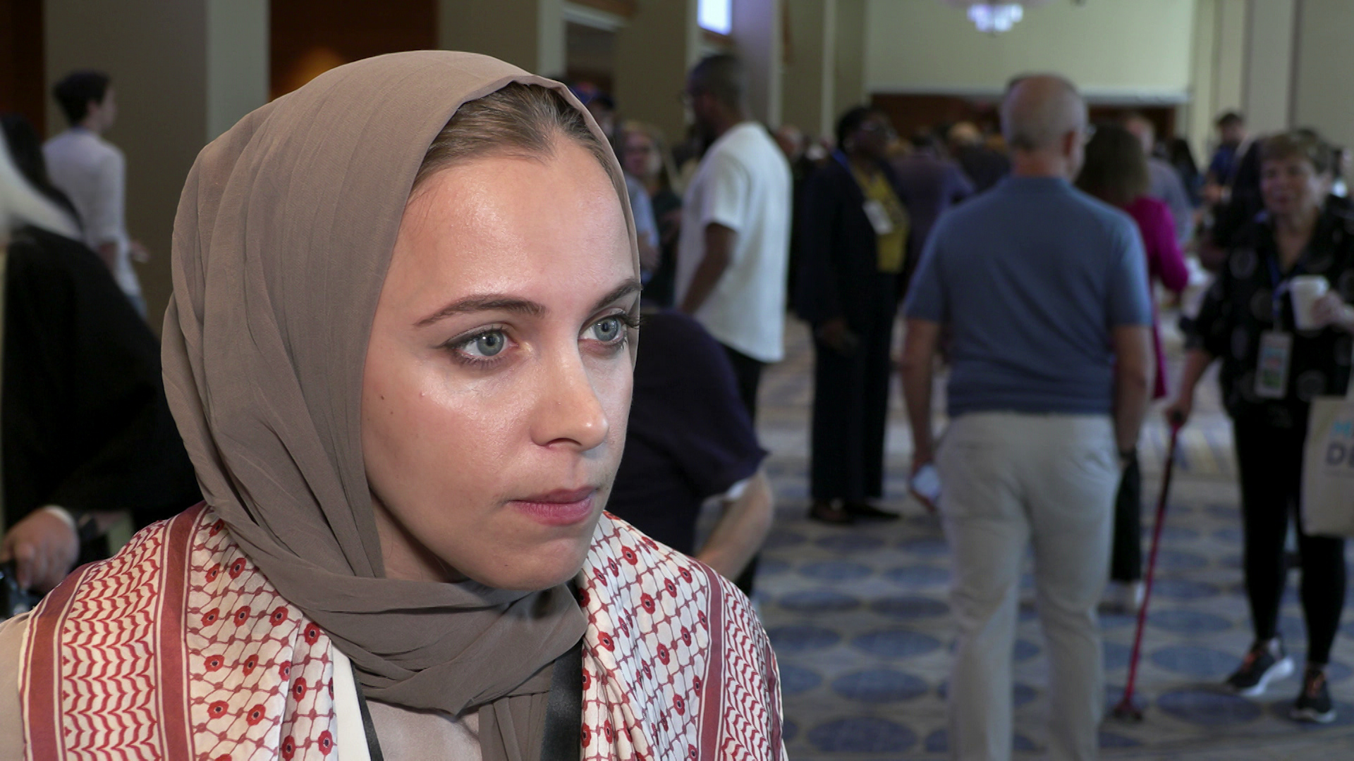 Halah Ahmad stands and speaks indoors with people walking in the background.