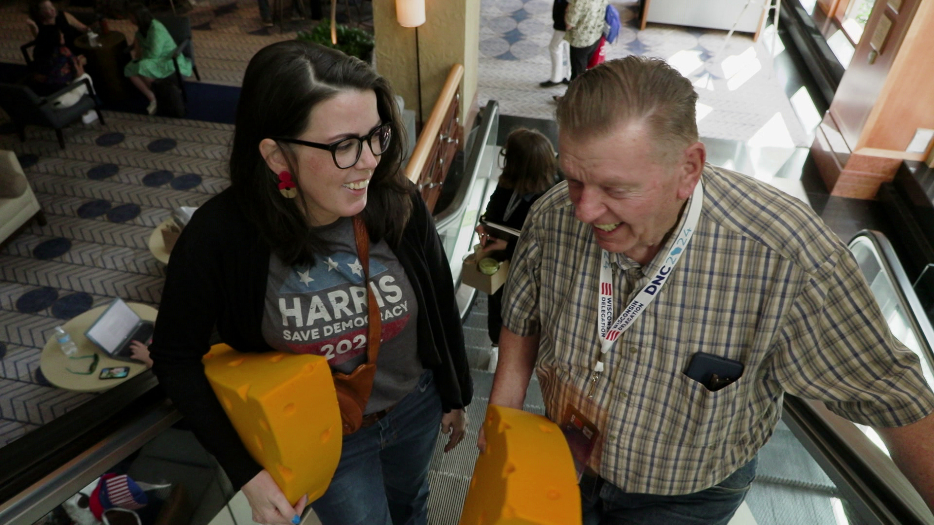 Gary and Kim Hawley hold foam cheeseheads while riding an escalator with people walking on the floor and tables below.