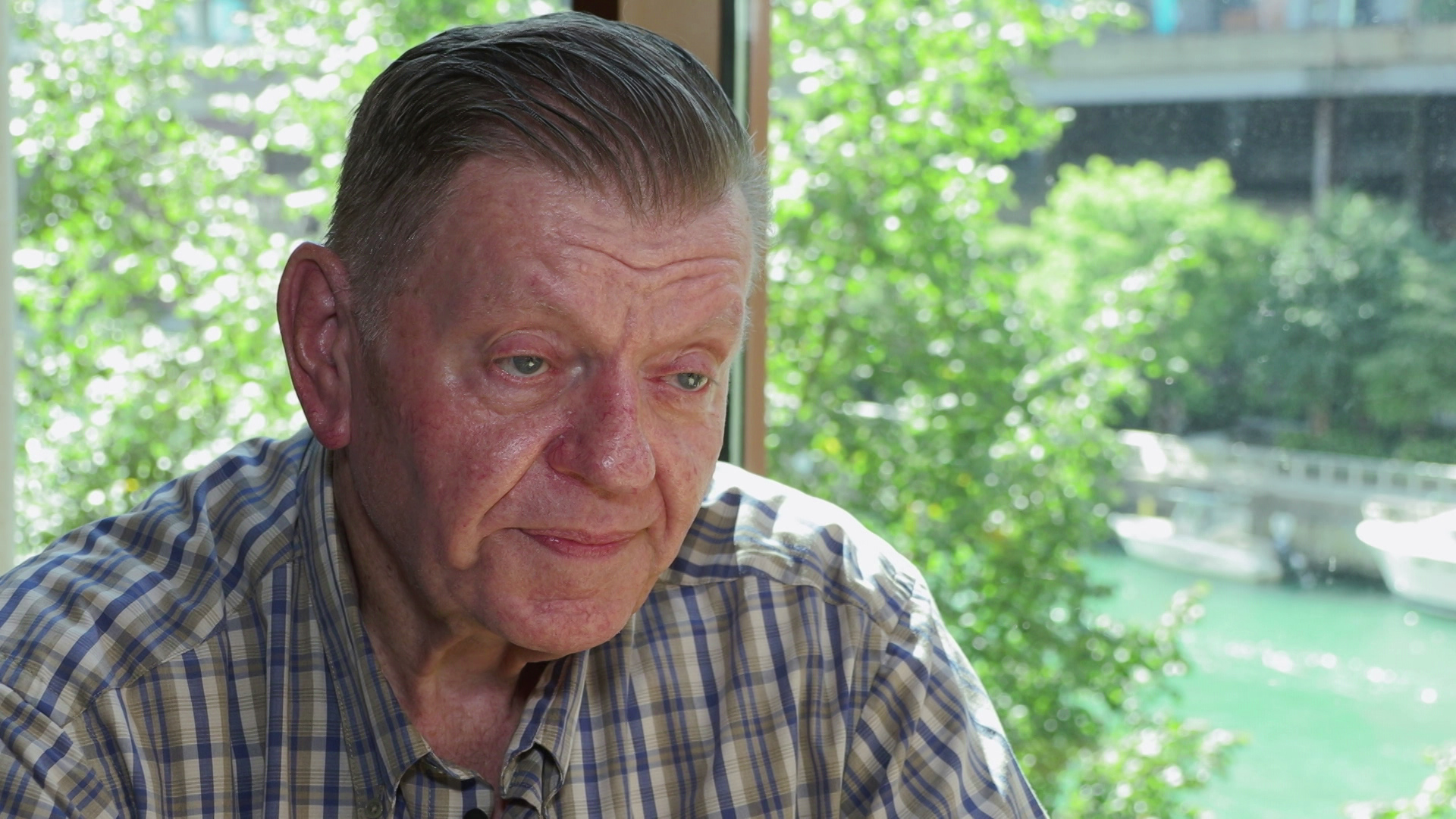 Gary Hawley sits and speaks in front of glass windows with trees, bushes and a building in the background.