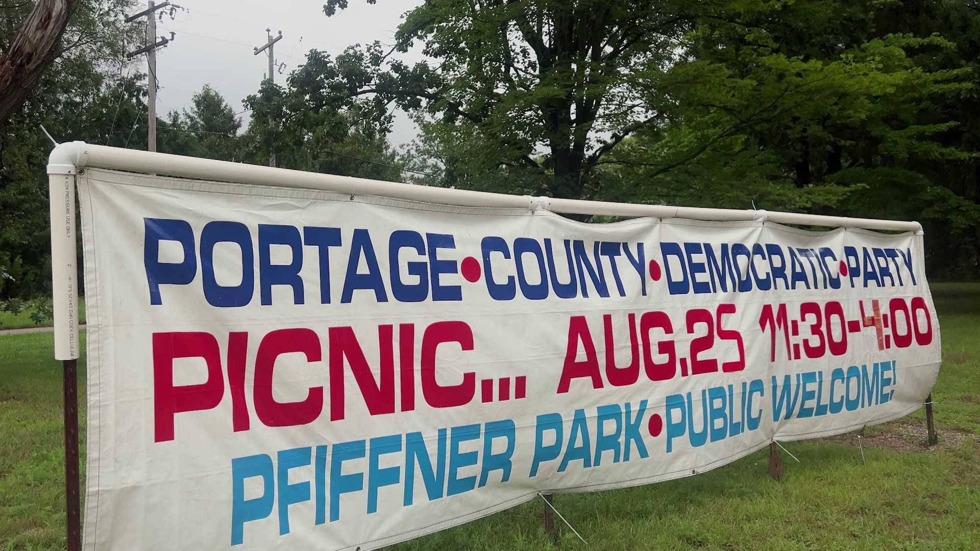 A sign reading "Portage County Democratic Party Picnic...Aug. 25 11:30-4:00 Pfiffner Park Public Welcome" is mounted on stakes and PVC pipe on a lawn with trees and power lines in the background.