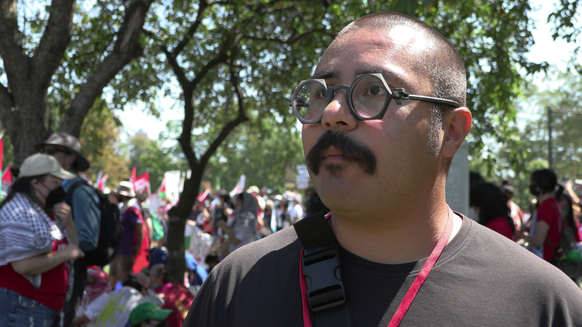 Omar Flores speaks while standing outside people walking and holding signs and flags amid trees in the background.