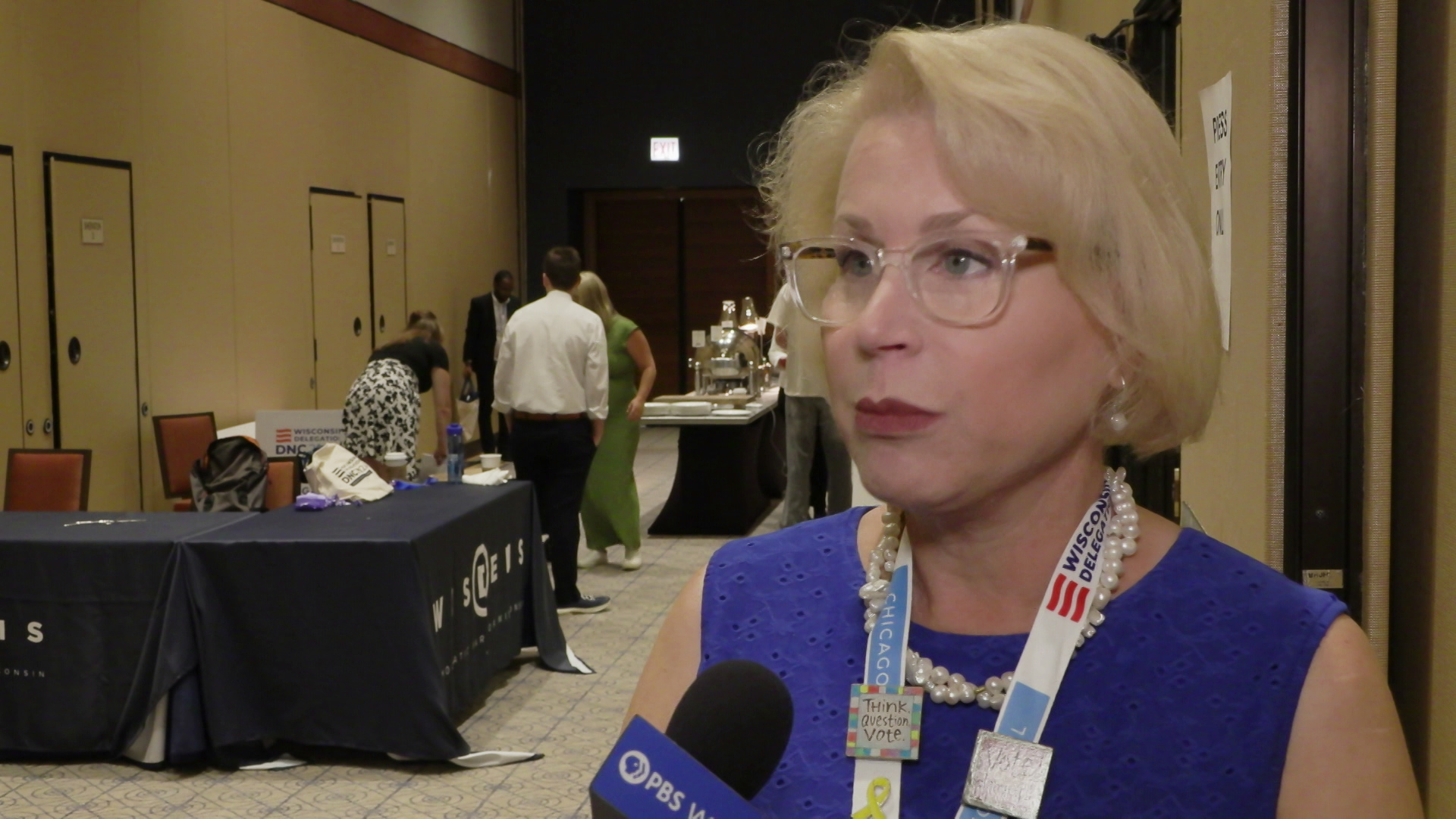 Ann Jacobs speaks into a microphone with a PBS Wisconsin flag while standing in a room with tables and people standing in the background.