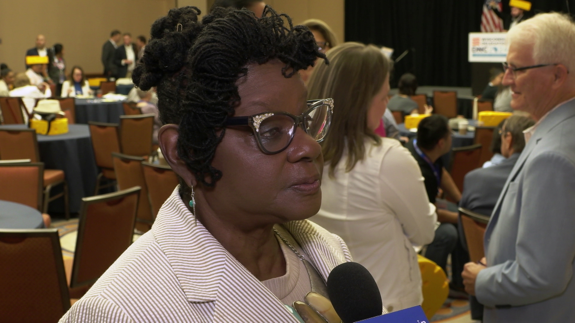 Gwen Moore stands and speaks in a room with rows of chairs and people standing and sitting in the background.