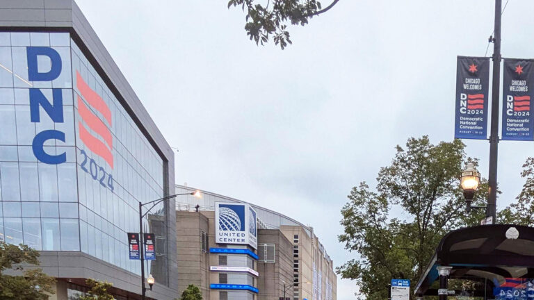 Vinyl signs with a six-pointed star and the words Chicago Welcomes above the wordmark for DNC 2024 are affixed to either side of light poles on a street, with a large version of the same logo on the corner a glass-walled building one one side of the street and a sign for the United Center next to an arena behind, with a bus shelter and trees on the opposite side of the street.