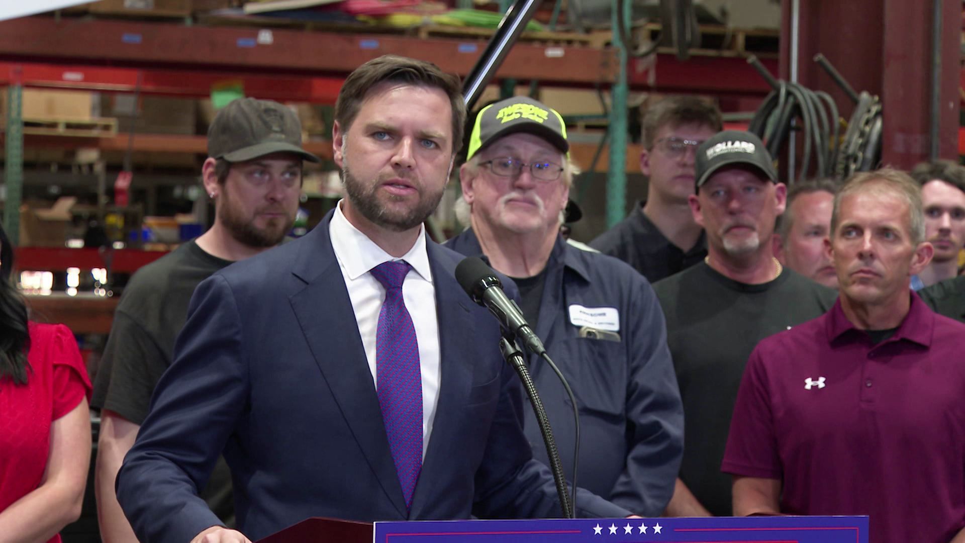 JD Vance stands and speaks behind a podium with a microphone in front of a group of people standing behind him with metal beams cables in the background.