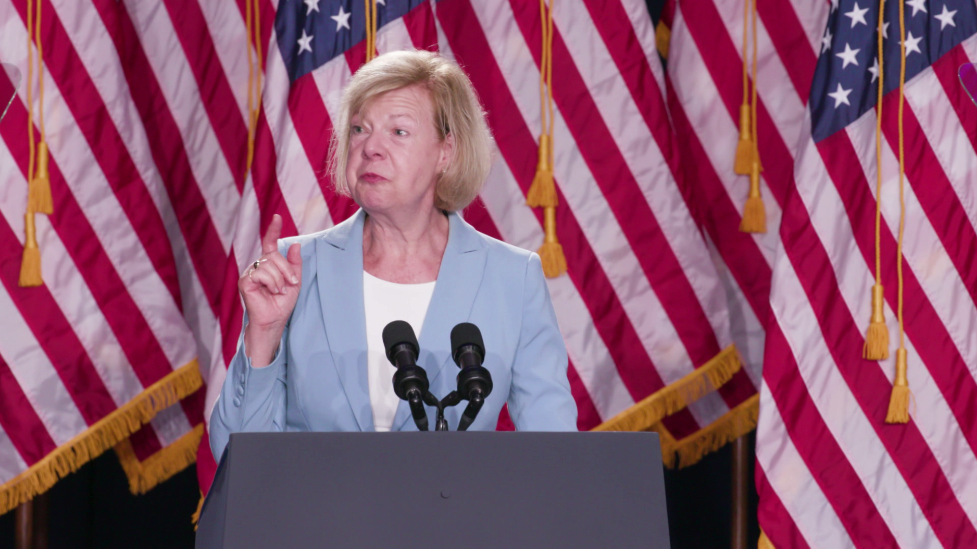 Tammy Baldwin stands and speaks behind a podium with two microphones and multiple U.S. flags in the background.