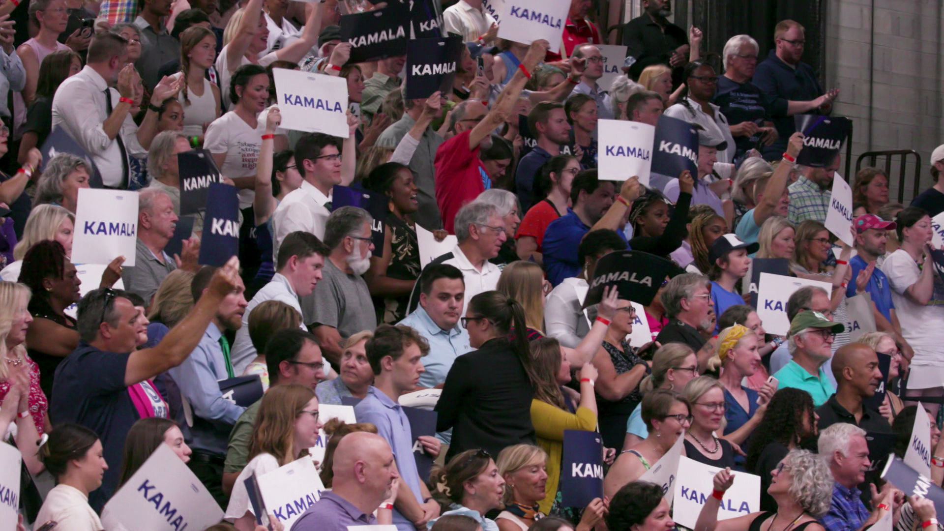 A crowd of people stand, cheer and hold signs that read 'Kamala' on bleachers in a gymnasium.