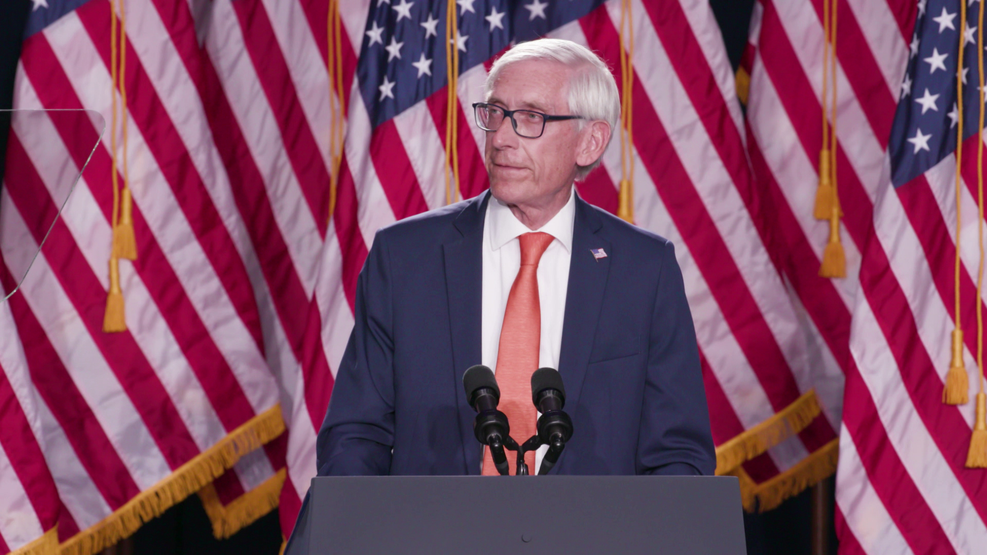 Tony Evers stands and speaks behind a podium with two microphones and multiple U.S. flags in the background.