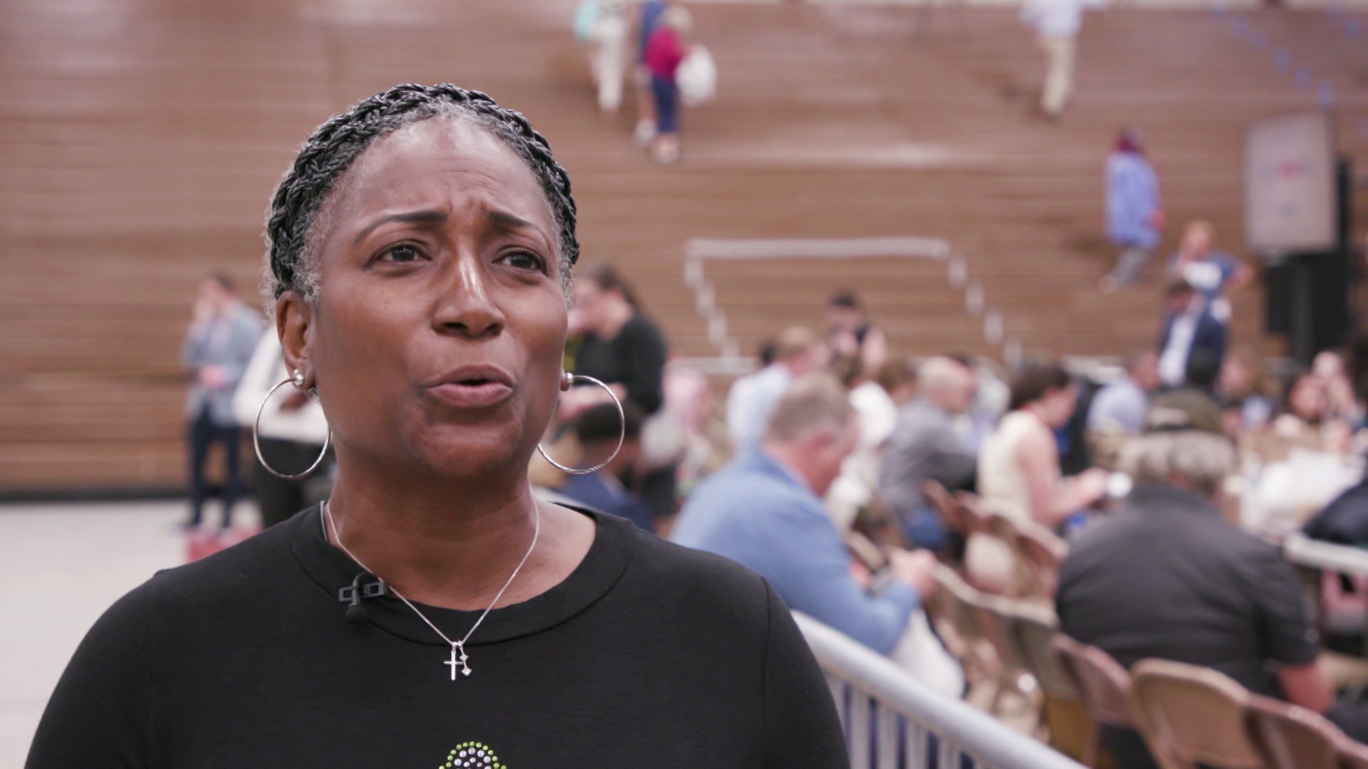 Julie Landry stands and speaks in a gymnasium with people seated and walking in the background.