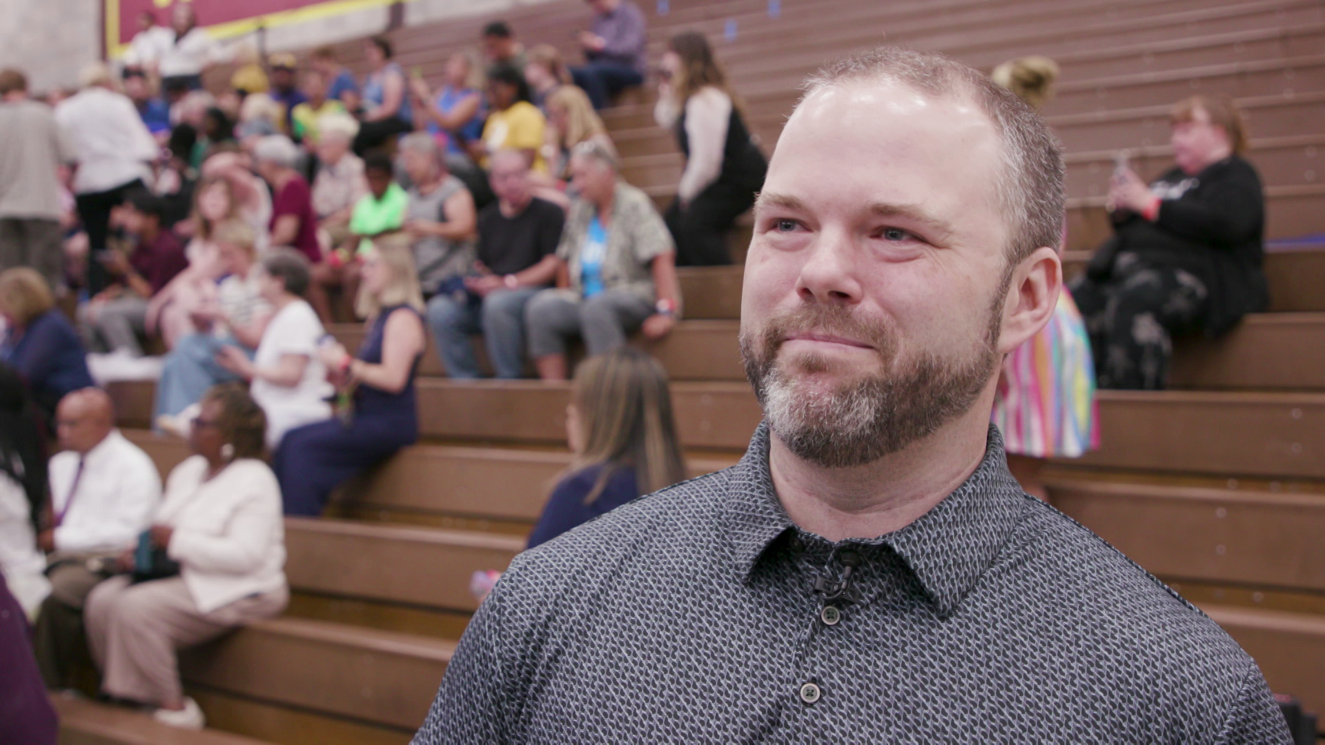 Paul McCreary stands and speaks in a gymnasium with people seated on bleachers in the background.