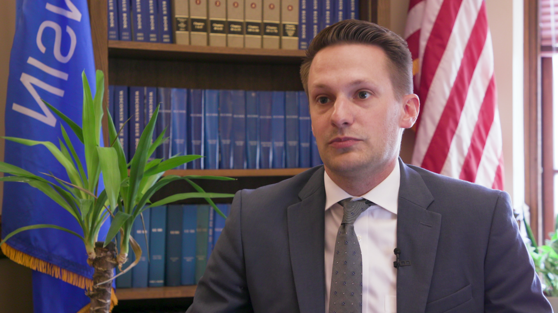 Alex Joers sits and speaks with a potted plant, bookshelf lined with books, and Wisconsin and U.S. flags in the background.